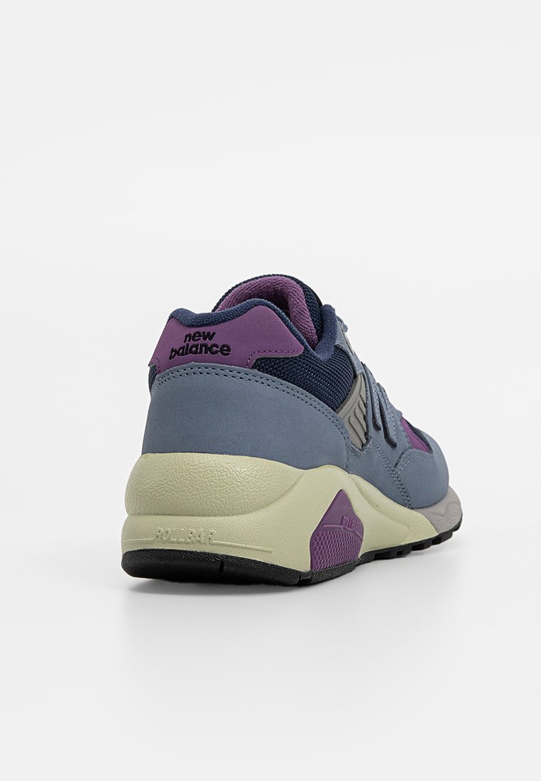 Zapatilla gris y marina con un acento púrpura, que presenta una parte superior texturizada, suela resistente y una marca visible en la lengüeta del talón.
