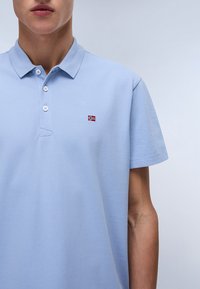 Svetlo modra polo majica iz teksturiranega materiala, z trojnim gumbom in majhno vezeno norveško zastavo na prsih.