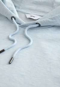 Sweat à capuche bleu clair avec cordons tressés et embouts métalliques, montrant l'étiquette intérieure du col avec la marque "NEXT".
