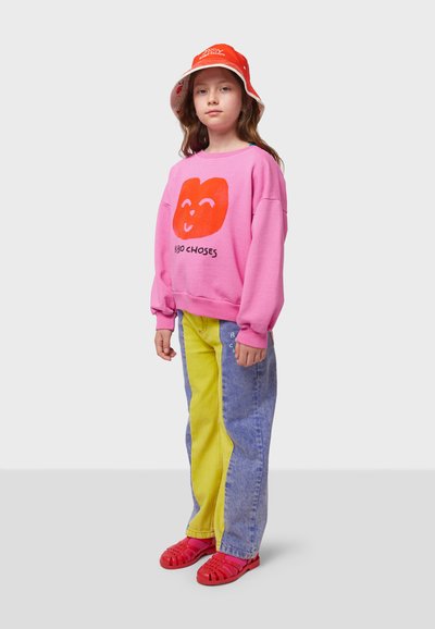 Pink sweatshirt with a red smiley face graphic, yellow and blue color-block denim pants, and red sandals. White background.