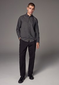 Camicia grigia con bottoni, maniche lunghe, pantaloni neri e stivali neri, con una lieve texture e vestibilità classica, su uno sfondo neutro.