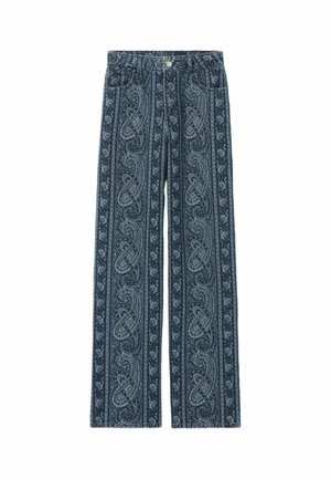 Femme portant une veste et un pantalon en jean bleu foncé à motif paisley assortis avec un chemisier bleu à motif paisley, debout avec une main dans la poche.