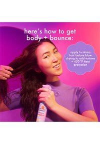 Produit capillaire dans une bouteille rose pastel et bleue avec des motifs colorés. Une personne tient la bouteille tout en coiffant des cheveux humides, vêtue d'un haut violet.