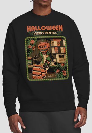 Man wearing black sweatshirt with Halloween video rental scene showing child returning a tape to a jack-o-lantern-headed clerk surrounded by pumpkins.
