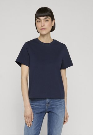 Femme aux cheveux courts et foncés, portant un t-shirt uni bleu marine à manches courtes et un jean bleu, debout devant un fond neutre.