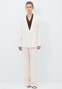 Krämfärgad kostym med smalrandigt mönster som har en skräddarsydd blazer med slagkrage och matchande vida byxor. Brun knapp-upplagd topp under.