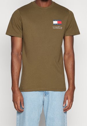 Hombre vistiendo una camiseta de manga corta de Tommy Jeans en color verde oliva con el logo en el lado izquierdo del pecho y jeans de color azul claro, de pie frente a un fondo liso.