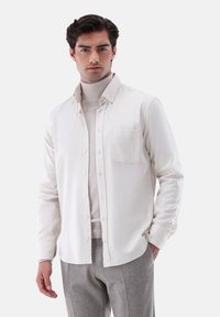 Weißes Hemd mit Knöpfen und einer einzelnen Brusttasche, kombiniert mit einem hellgrauen Rollkragenpullover. Glatter Stoff, taillierte Passform, minimales Design.