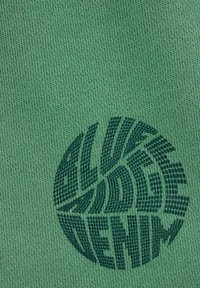 Zeleno blago z teksturirano pleteno vzorcem, ki vsebuje temnozeleni krožni logotip z napisom "ALIVE DENIM." Logotip ima obliko lista.