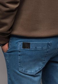 Jasnoniebieskie dżinsy z teksturowaną, czarną skórzaną naszywką na tylnej kieszeni. Miękka tkanina o dopasowanym kroju, z wzmocnionymi szwami.