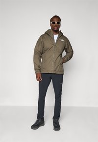 Veste à fermeture éclair verte olive avec capuche, associée à un pantalon noir et des chaussures de randonnée noires. Logo notable sur la veste. Lunettes de soleil grises portées.
