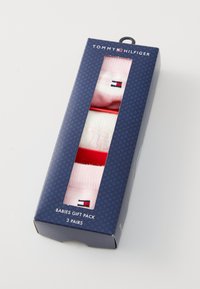 Darilni paket za dojenčke, ki vsebuje tri pare nogavic iz svetlo rožnate tkanine, vsake nogavice pa krasijo majhne oznake v rdeči, beli in modri barvi.