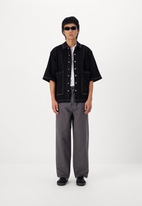 Svart denim kortermet skjorte med hvite sømmer, kombinert med grå løstsittende bukser og svarte sneakers. Tilbehøret inkluderer mørke solbriller.
