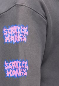 Grauer Sweatshirt mit zwei großen Logos. Die Logos haben fett gedruckten, gezackten pinken und blauen Text mit der Aufschrift "SERVICE WORKS". Die Textur wirkt weich.