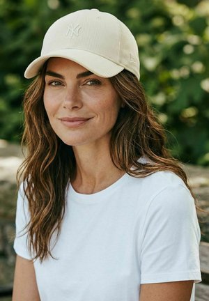 Woman with wavy brown hair wearing a white NY baseball cap and white t-shirt, smiling softly against a blurred green outdoor background.