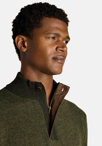 Noor koristav noormees, kellel on lühikesed lokkis juuksed, kannab oliivroheline tõmbluku villajakki pruunist nubukist kraega, vaadates paremale.