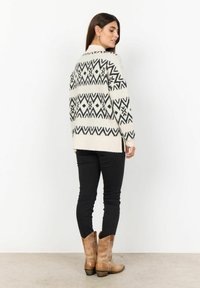 Knitted sweater in cream with black geometric patterns, long sleeves, and side slits. Paired with slim black pants and brown ankle boots.