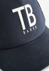 Kapa temno modre barve iz teksturirane tkanine, s prominentnim belim logotipom "TB" in napisom "BAKER", strukturna zasnova ter ukrivljenim robom.