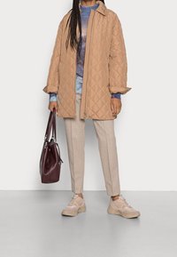 Personne portant un manteau matelassé beige, un pullover tie-dye, un pantalon beige et des baskets, tenant un grand sac à main en cuir rouge foncé.