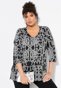 Tunic nera con un motivo geometrico bianco, scollo a barca, maniche a tre quarti e spacchi laterali. La modella tiene una pochette argento.