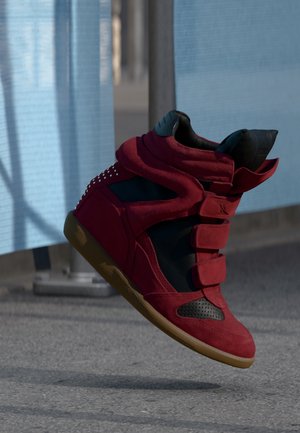 Czerwono-czarny but sportowy typu high-top z dwoma paskami i gumową podeszwą uniesiony nad szarym chodnikiem przy metalowej barierce z niebieską tkaniną.