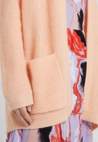 Light peach knitted cardigan with ribbed texture, open front, and a front pocket, layered over a multicolored abstract patterned dress.