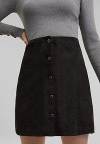 Zwarte suède mini-jurk met zes bruine knopen aan de voorkant, met een gladde textuur en een getailleerd ontwerp. Bovenaan een grijze ribbeltrui.