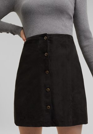 Mini jupe en suedine noire avec six boutons marron sur le devant, présentant une texture lisse et un design ajusté. Pull gris à côtes au-dessus.