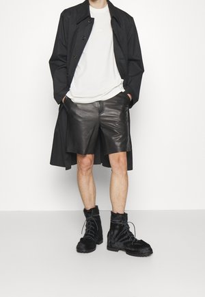 Czarny płaszcz trench na białej koszulce, w zestawie z czarnymi skórzanymi szortami i czarnymi botkami z futrem. Gładka faktura, użyteczny design.