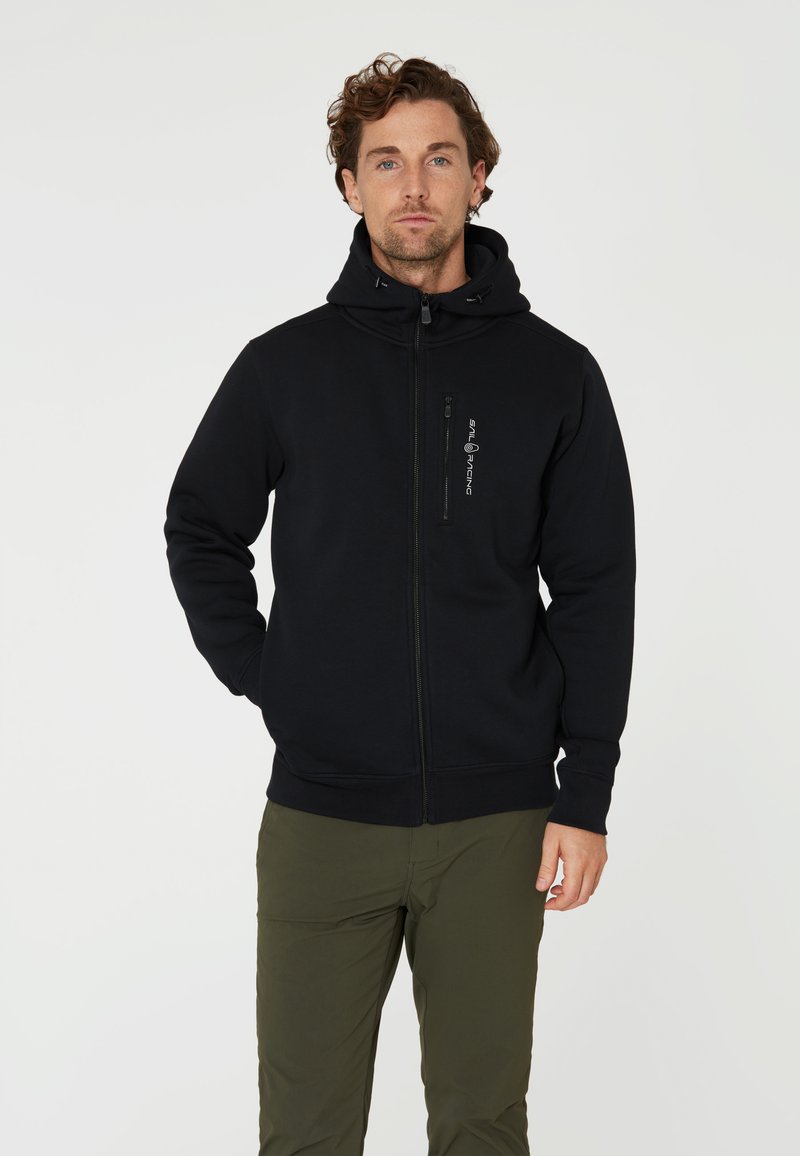 Svart zip hoodie i mjukt material, med hög krage och en logotyp på bröstet. Ribbad manschetter och nederkant. Bärs med olivgröna byxor.
