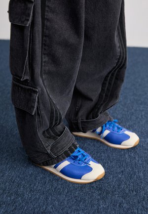 Persoon in losse zwarte cargobroeken en blauw-witte sneakers staand op een donkerblauwe vloer.