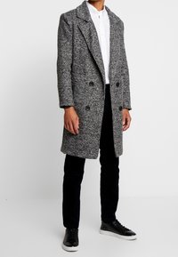 Šedý texturovaný dvouřadový kabát s velkými černými knoflíky, klopovými límci a dlouhými rukávy, nošený přes bílou košili a tmavé kalhoty.