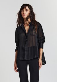 Frau mit langen braunen Haaren, die eine durchsichtige schwarze Bluse mit Knöpfen und schwarze Hosen trägt, vor einem schlichten hellen Hintergrund.