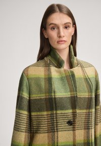 Grüner und beiger karierter Mantel aus Wollmischgewebe mit hohem Kragen und Ein-Knopf-Verschluss, der ein strukturiertes Fischgrätmuster zeigt.