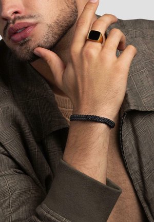Black beaded bracelet on wrist, gold square ring on finger, and a gray plaid jacket with a brown textured sweater underneath.