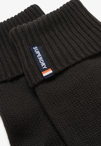 Sort strikket sweater med ribbet manchet. Har et stofmærke med "SUPERDRY" i hvidt på en navy baggrund, pyntet med røde og hvide striber.