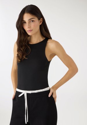 Kobieta o długich ciemnych włosach, ubrana w czarną koszulkę bez rękawów i czarne spodnie z białym paskiem ze sznurkiem, stojąca na jednolitym tle.