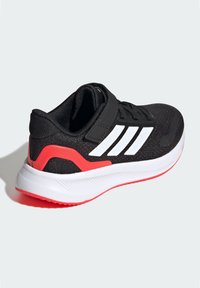 Chaussure de sport noire avec une tige en mesh, des rayures blanches et un détail de talon rouge ; dispose d'un col rembourré et d'une sangle à boucle ajustable.