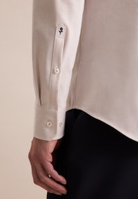 Lichtbeige langemouwshirt van een textuurstof, met een zwarte geborduurde roos op de manchetten en witte knoopsluitingen.