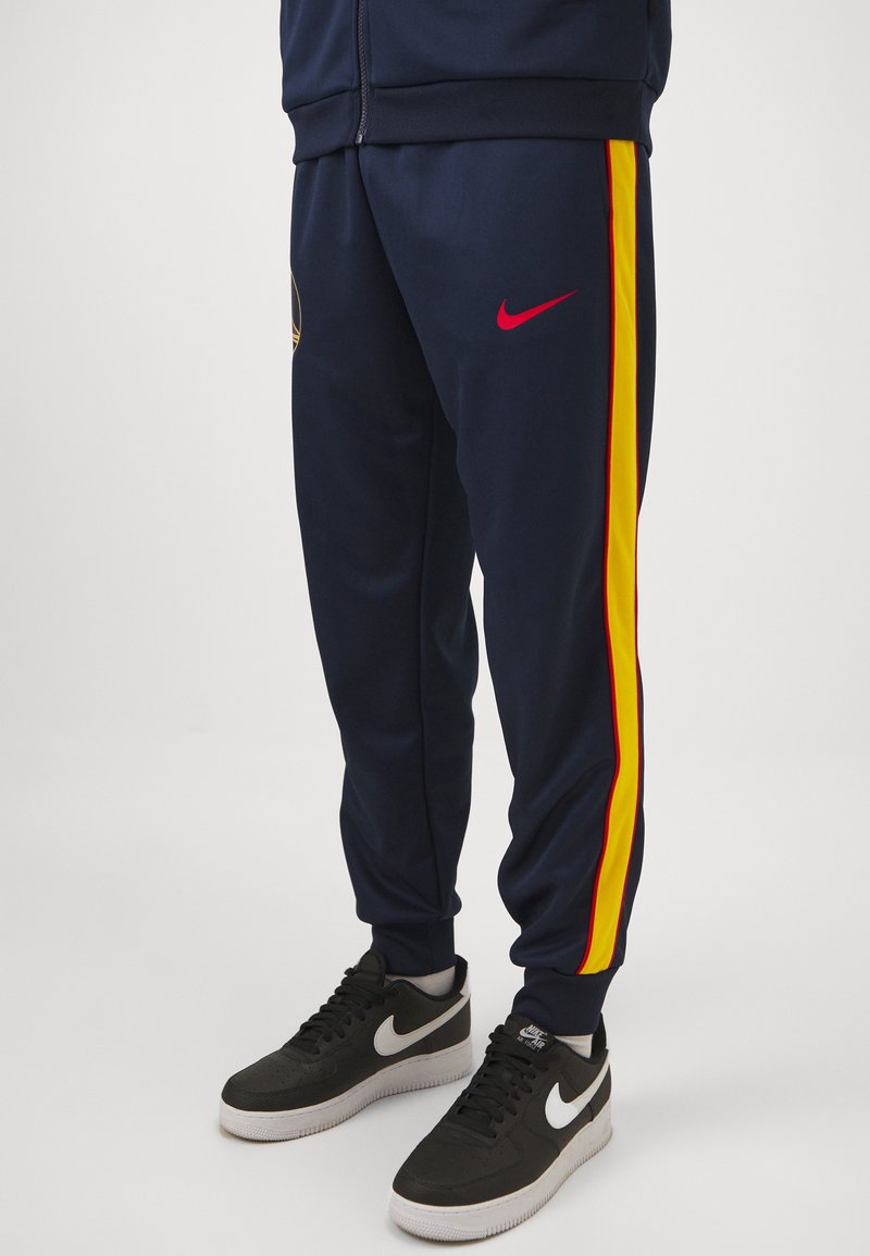 Pantaloni sportivi blu navy con strisce laterali gialle e rosse, polsini elastici e logo Nike rosso. Indossati con scarpe da ginnastica nere Nike.