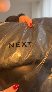 Juoda plastikinė pakuotė su žodžiu „NEXT“ parašytu ryškiais, kontrastingais didžiosiomis raidėmis; laikoma rankoje, kurios nagai nudažyti raudonai.