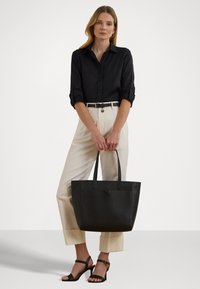 Black leather tote bag with embossed logo, smooth texture, structured shape, and double handles. Paired with a black shirt and cream trousers.