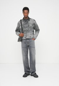 Pilkas džinsinis švarkas su priekinėmis kišenėmis, derinamas su laisvo kirpimo pilkais džinsais, pagražintais mėlynomis siūlėmis. Modelis laiko juodą per petį nešiojamą krepšį.