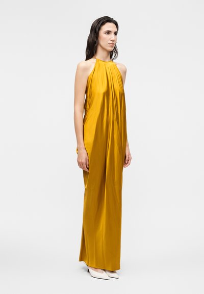 Γυναίκα με μακρύ, μουσταρδί φόρεμα halter neck και άσπρα μυτερά ψηλοτάκουνα παπούτσια, στέκεται μπροστά σε ένα απλό ανοιχτό γκρι φόντο.