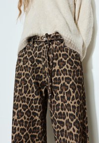 Kobieta w beżowym, miękkim swetrze włożonym do wysokich, szerokich spodni w panterkę z paskiem zawiązanym w talii.