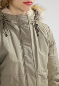 Žena v béžovém zimním parku se šedým kožíškem a vlněnou podšívkou, odhalující krk a červené rty na jednobarevném pozadí.