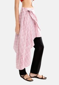 Różowa spódnica w kratkę o drapowanej konstrukcji, związana w talii, zestawiona z czarnymi spodniami i brązowymi sandałami na paski.