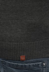 Pull en tricot gris foncé avec un ourlet côtelé et une petite étiquette en cuir en bas. Il est associé à un jean en denim bleu.