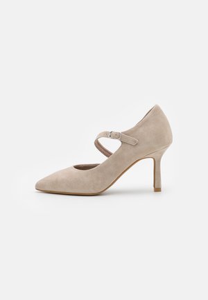 Beige suède Mary Jane-hakken met een spitse neus, verstelbare band en slanke hak op een effen witte achtergrond.