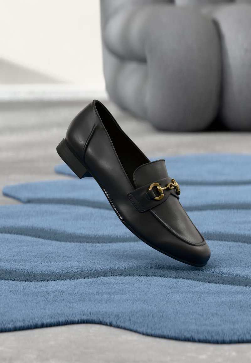 Mocassin en cuir noir avec détail de boucle dorée posé sur un tapis bleu texturé, avec en arrière-plan une sculpture grise floue.